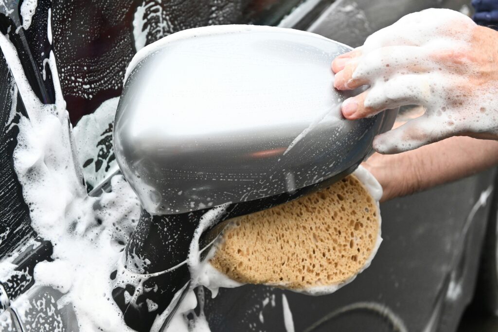 Home 5 A person washes a car mirror with a soapy sponge, showcasing foam and water.